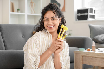 Beautiful young happy African-American woman with makeup brushes doing makeup at home