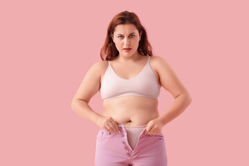 Overweight young worried woman in tight jeans on pink background. Weight gain concept
