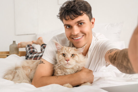 Handsome young happy man with cute Maine Coon cat taking selfie in bedroom at home