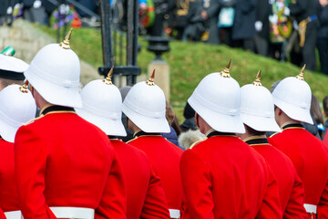A group of men wearing bright red uniforms with wide white belts standing outside at a parade ceremony. The young military soldiers have white helmets with gold metal spikes or bonnets. 