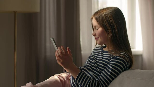 Caucasian happy little girl lying on sofa holding mobile phone smartphone cellphone gadget child kid daughter technology texting chat online pleasure smile home connect internet sms communicate female