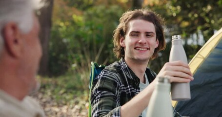 Camping, man and father toast in nature on outdoor holiday vacation for cheers, family or natural forest. Flask bottle, coffee and happy senior dad drinking with son on adventure in Australia woods