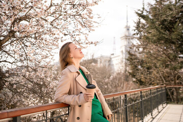 A woman is standing on a ledge with a cup of coffee in her hand. She is wearing sunglasses and a...