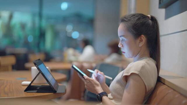 A woman is sitting at a table with a tablet and a cell phone in front of her. She is using a stylus to write on the tablet. The scene takes place in a cafe, with other people visible in the background