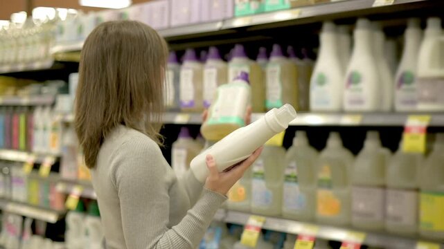 A person examining a bottle of laundry detergent in a supermarket aisle, considering options among a variety of cleaning products. High quality 4k footage