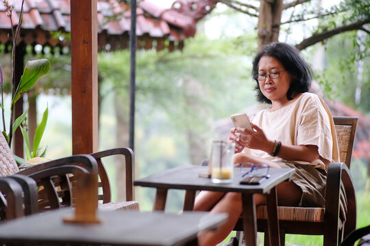 Middle aged Asian woman sitting at her home veranda, reading messages on her smartphone