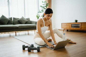 Young Asian woman sitting on the floor on yoga mat in front of laptop and training at home. Beautiful smiling woman doing stretching exercise while communicates with a trainer online before workout