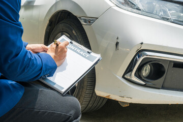 A businessman stands next to his damaged vehicle after an accident, holding a clipboard and...