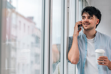 young man with mobile phone and cup at home window