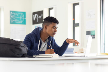 In school, African American male student using laptop and writing in notebook, studying