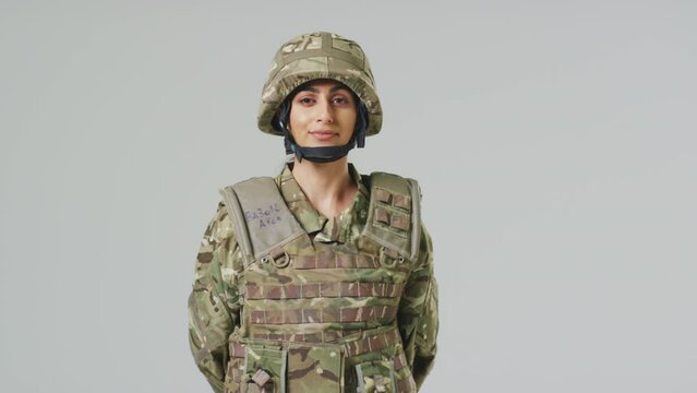 Young female soldier in uniform wearing body armour and helmet in front of plain studio background - shot in slow motion