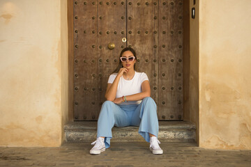 Young beautiful woman with brown hair, sunglasses, white t-shirt and blue pants, posing sitting on...