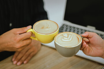Asian man and woman, enjoy a morning meeting at a cafe, holding cups of hot lattes, clinking together in a cheerful, relaxed atmosphere with a blurred background.