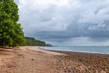 A curved beach under glowering overcast skies with tropical rainforest growing up to the edge of the beach which is strewn with stones exposed by the tide at Mission Beach in Queensland, Australia.