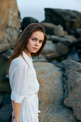 Serene young woman in flowing white dress standing on ocean rocks by the shore