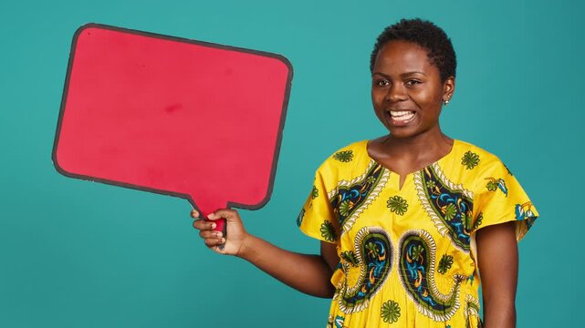Smiling person showing a red isolated speech bubble in studio, holding a cardboard icon for a commercial advertisement. Young woman in tribal attire presenting an empty carton sign. Camera A.