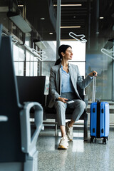 Business woman sitting in airport and waiting for starting her travel voyage. Woman with luggage is holding handle of the suitcase and looking around airport terminal.
