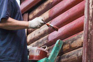 A man works with a brush and applies red paint to a wooden village house. A worker paints wooden planks in close-up. Half of the painted surface. Smearing with a brush.