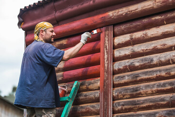 A man works with a brush and applies red paint to a wooden village house. A worker paints wooden planks in close-up. Half of the painted surface. Smearing with a brush.