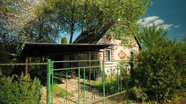 Footage of a brickstone summer house with a garden and a green iron gate swaying in the wind during spring. The scene highlights the charm and tranquility of the countryside. Location: Baltics
