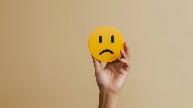 Hand Holding a Sad Emoji on a Beige Background suitable for business background, posters, wallpapers, banners, greeting cards, and advertising for business entities or brands.