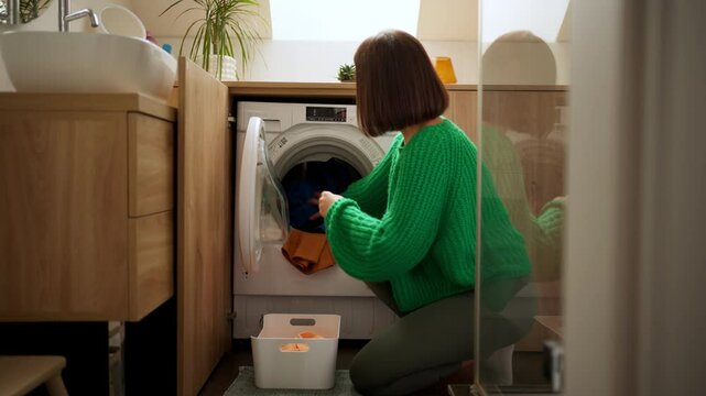 Young housewife opening door of washing machine and putting dirty clothes laundry inside at home