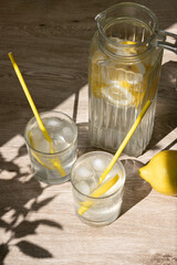 Fototapeta premium Two glasses and jug with lemonade drink on beige wooden table background with aesthetic natural floral sunlight shadows