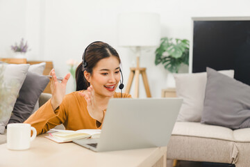 Naklejka premium Smiling woman wearing a headset, engaged in video call while working from home with laptop and taking notes.