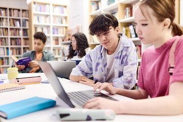 Portrait of young teenage boy looking at laptop screen in school library with group of students copy space