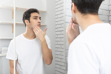 Handsome man looking at mirror and applying moisturizing cream on cheeks in bathroom. Groomed young guy doing skincare morning routine after taking a shower