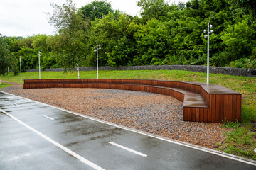 A quaint curved wooden bench sits by the roadside, enhancing the urban scenery with its charm. The street is bordered by lush greenery and city buildings, creating a picturesque view