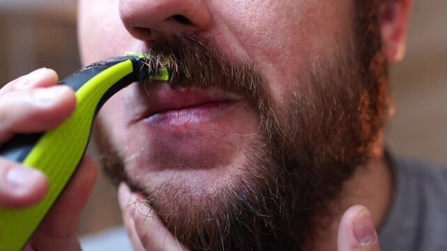 The man is using a trimmer to shave his beard. He carefully maneuvers around his jaw and mouth, being cautious not to nick his skin with each movement of his hand and fingers