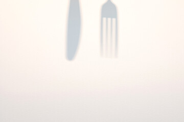 Close-up image of shadows cast by a knife and fork on a white paper texture. Food menu copy space background.