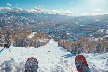 A skier's perspective of a snowy mountain slope, with ski tips visible in the foreground. Landscape features snow-covered trees, rolling hills, and clear blue skies