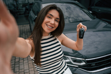 Making selfie, holding keys, happy owner. Attractive young woman is outdoors near her brand new car