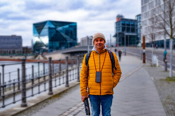 Cheerful middle-aged man, 65 in winter bright yellow puffer jacket and beanie, smiling on urban street with historic buildings in background