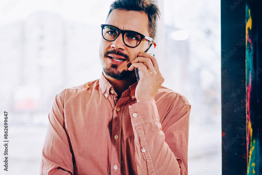 Poster male businessman making dissatisfied face while talking with service operator on mobile phone, young