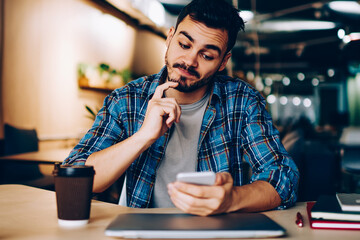 Contemplative male analyzing information about booking on website using cellular in coworking...