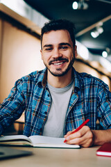 Portrait of cheerful male student enjoying learning in coworking office using laptop computer for research,happy freelancer looking at camera during making project for remote job making notes