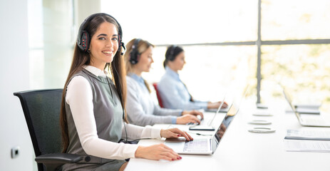 Young woman wearing a headset is sitting at a desk with a laptop, with her colleagues seated in a row in the background.