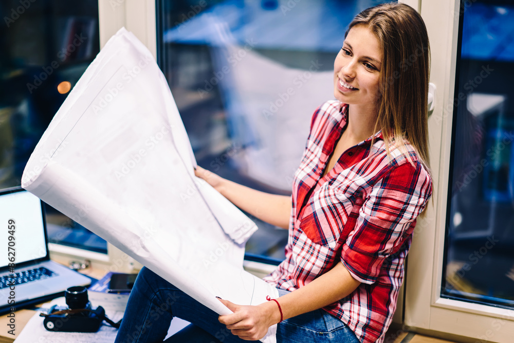 Poster smiling woman holding papers with drawings satisfied with completing blueprint before deadline sitti