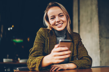 Half length portrait of happy female blogger dressed in stylish apparel updating profile on smartphone device with 4G internet connection while smiling at camera.Cheerful owner with mobile in hands