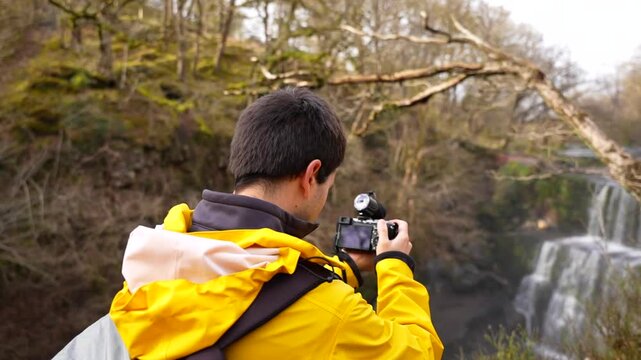 Man in a yellow jacket adjusting his camera lens to photograph Sgwd Isaf Clun-Gwyn Waterfall, Wales