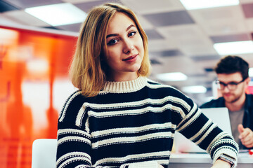 Portrait of charming blonde young woman dressed in casual outfit looking at camera while sitting in office interior.Attractive student with smartphone in hand during break in univerisity