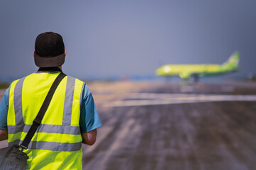 Young man in yellow safety vest observing airplane on airport runway. Camera case on shoulder. Aviation dream concept. Blurred background. Back view