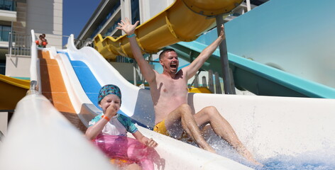 Obraz premium Joyful dad and daughter go down slide in water park. Summer vacation and vacation with children concept