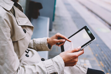 Side cropped view of young woman with finger touching on blank copy space display of modern tablet to make payment online on website via free 4G internet connection sitting outdoors on tram stop