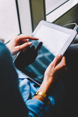 Cropped back view of young woman holding modern tablet in hands and touching with finger on display using free transport wifi in tram.Female passenger in earphpnes choosing music app on touch pad
