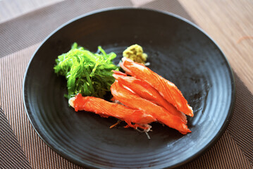 Close up crab sticks sashimi or Kani sashimi in a small black plate with wasabi and seaweed. Natural light from window, view from above around 45 degree.