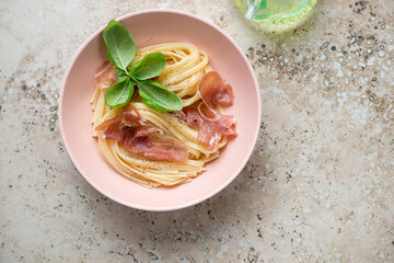 Linguine with prosciutto and green basil in a roseate bowl, top view on a beige granite background, horizontal shot with space